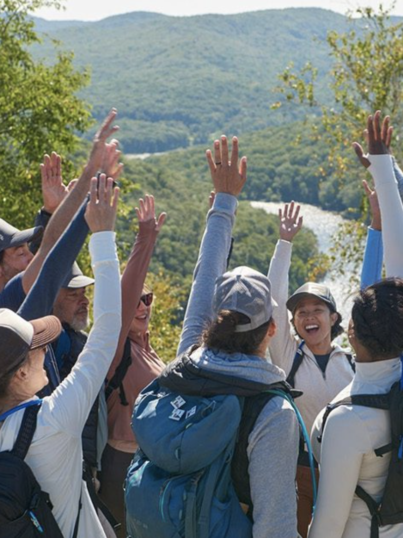 Group of hikers in outdoor gear standing in a circle, raising their hands in cheer, with a scenic woodland and mountain view in the background.