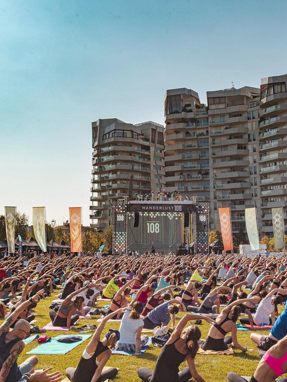 A large group of people practice yoga outdoors on a grassy field in front of modern high-rise buildings. A large screen displaying "108" is visible in the background.