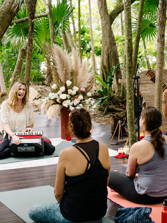 A group of people participate in an outdoor yoga session in a forested area, with a woman playing a keyboard and a man playing a sitar at the front of the class.