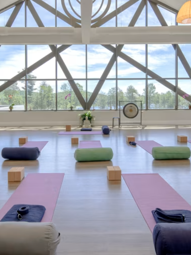 A spacious yoga studio with large windows. Several yoga mats, bolsters, blocks, and blankets are neatly arranged on the floor. A gong stands in the back of the room under a geometric window design.