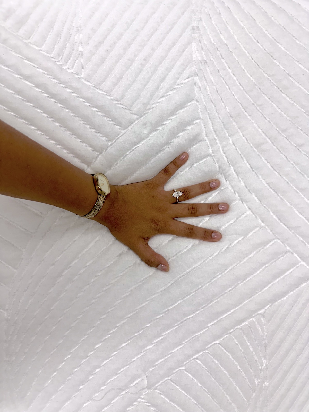 A hand wearing a watch and a ring is pressed against an organic mattress with a white textured surface.