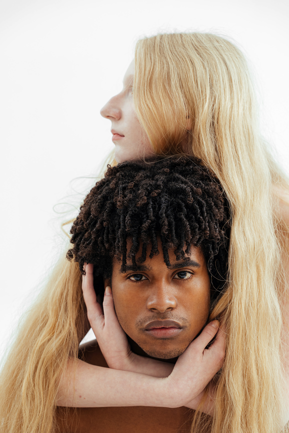 A person with long blonde hair embraces another person with short, curly hair from behind, both gazing forward with neutral expressions against a plain white background.