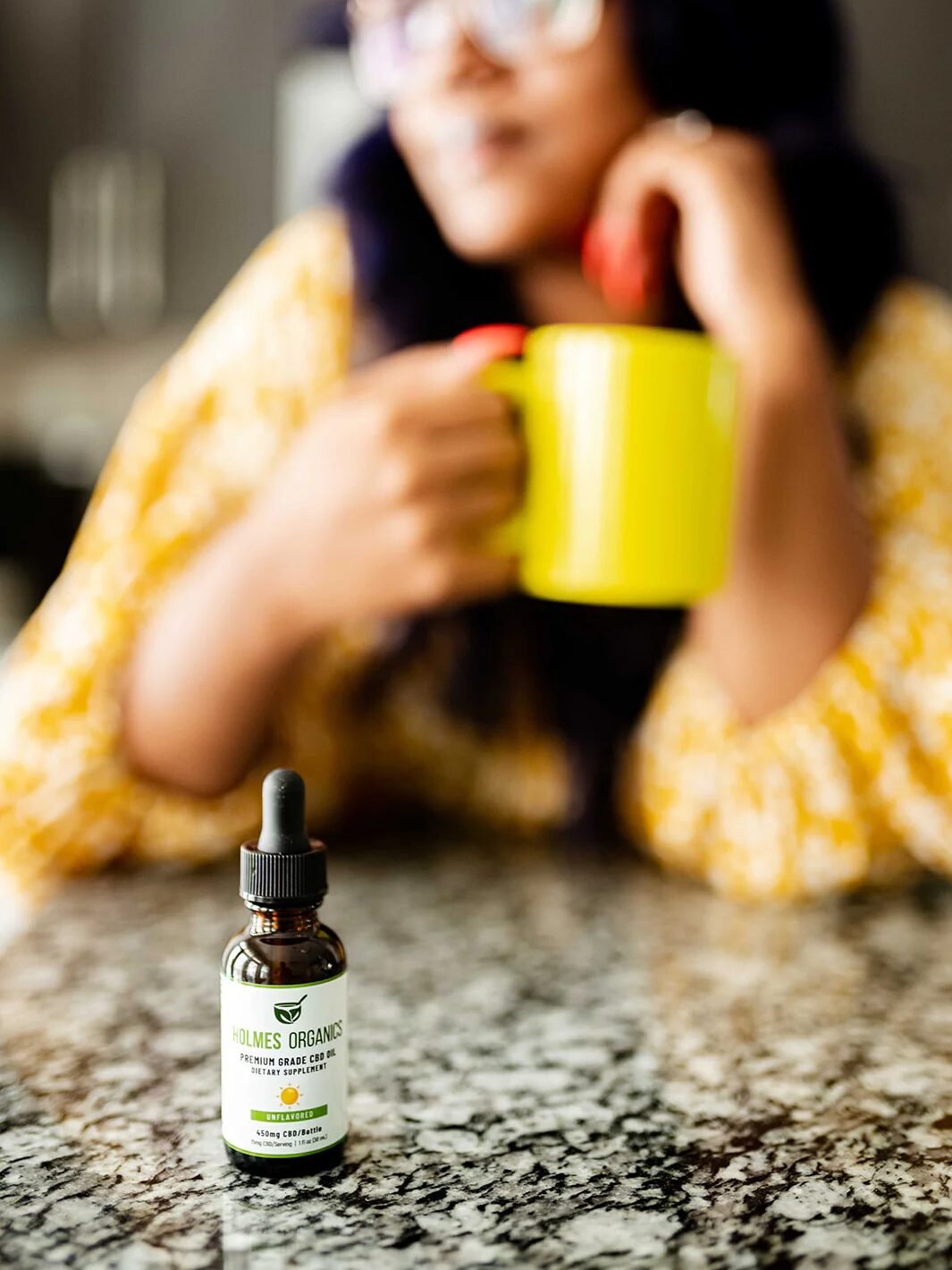 A person holding a yellow mug is blurred in the background, with a bottle of Holmes Organics CBD oil placed on a granite countertop in focus in the foreground.