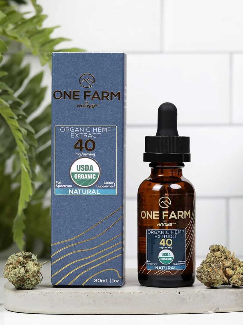 A 30ml bottle of One Farm Organic Hemp Extract with a dropper sits next to its blue boxed packaging. The label shows it contains 40 mg per serving and is USDA organic. Several hemp buds are placed nearby.