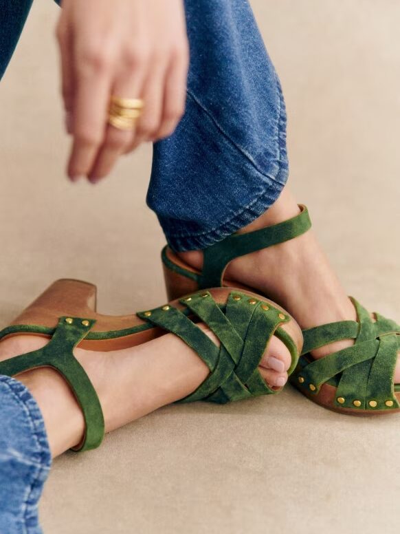 A person wearing green strappy sandals with gold studs, paired with blue jeans. The person's hand with gold rings is resting on their knee.