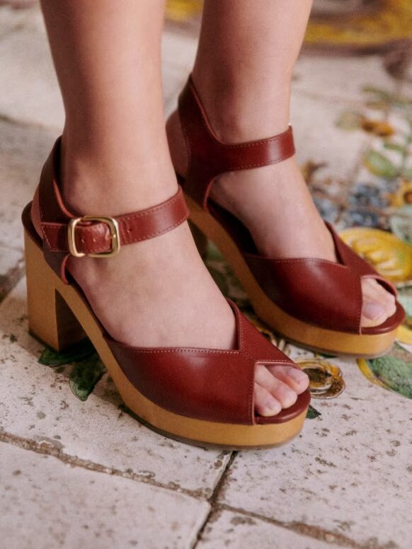 A close-up image of a person wearing brown leather high-heeled sandals with ankle straps and buckles, standing on a tiled floor.