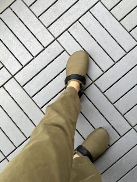 Person wearing tan pants and beige shoes with black straps standing on a gray tiled floor arranged in a diagonal pattern.