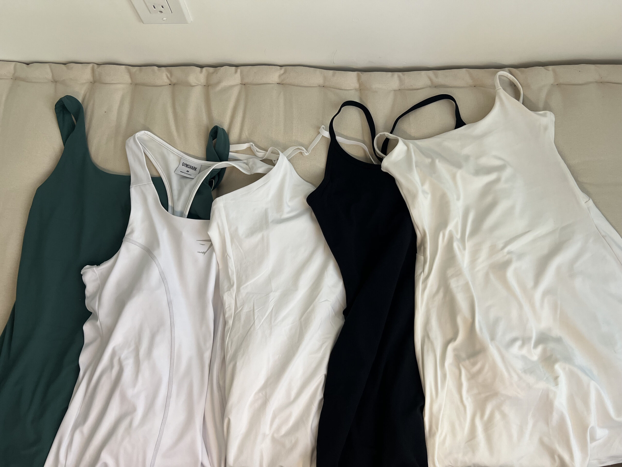 Five tank tops in various colors (green, white, black, and off-white) are laid out on a beige surface, arranged side by side.