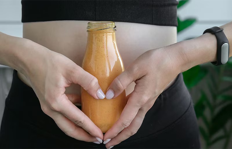 A person wearing a black fitness outfit holds a glass bottle of orange juice, ideal for pairing with fiber supplements for women, in front of their midsection. A fitness tracker is on their right wrist.