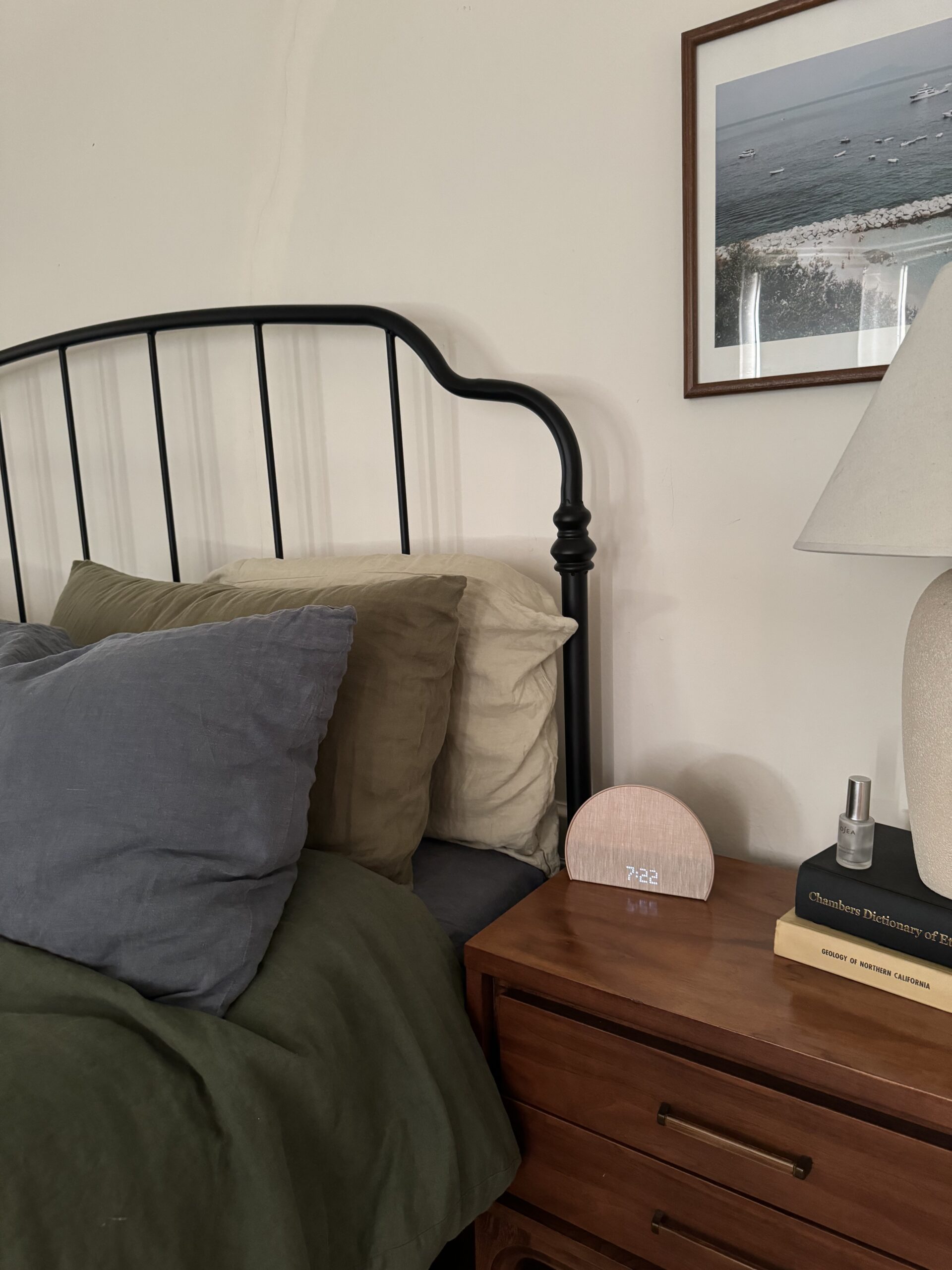 A bedroom with a black metal headboard, green and gray bedding, a wooden nightstand with a lamp, books, and a clock displaying 7:22. A framed photo hangs on the wall above the nightstand.