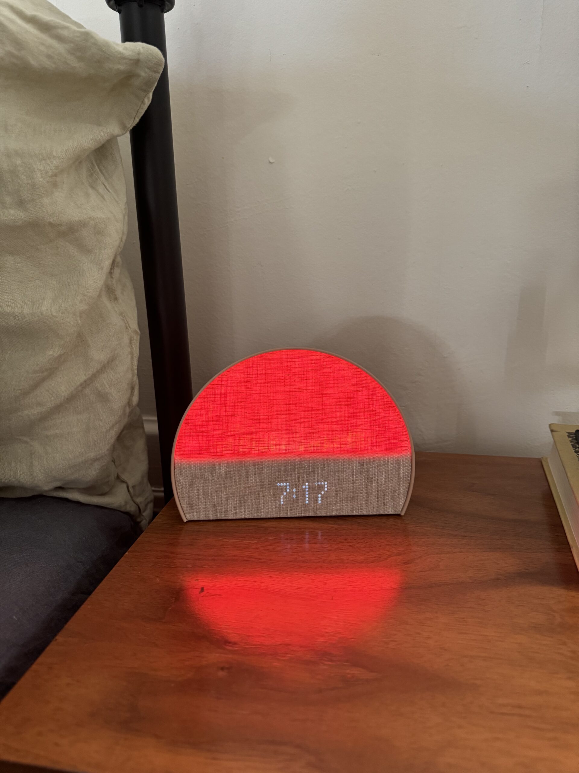 A digital alarm clock displaying 7:17 is on a wooden nightstand. The clock emits a red light and is positioned next to a bed with a white pillow and a stack of books.