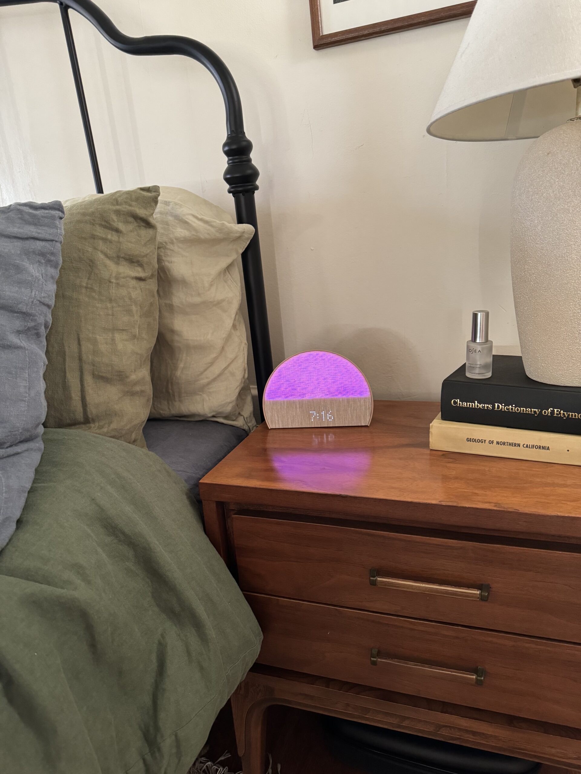 A nightstand with a wooden drawer cabinet, a clock showing 7:16 with a purple light, a book, a lamp, and a bottle, situated next to a bed with green and beige bedding.