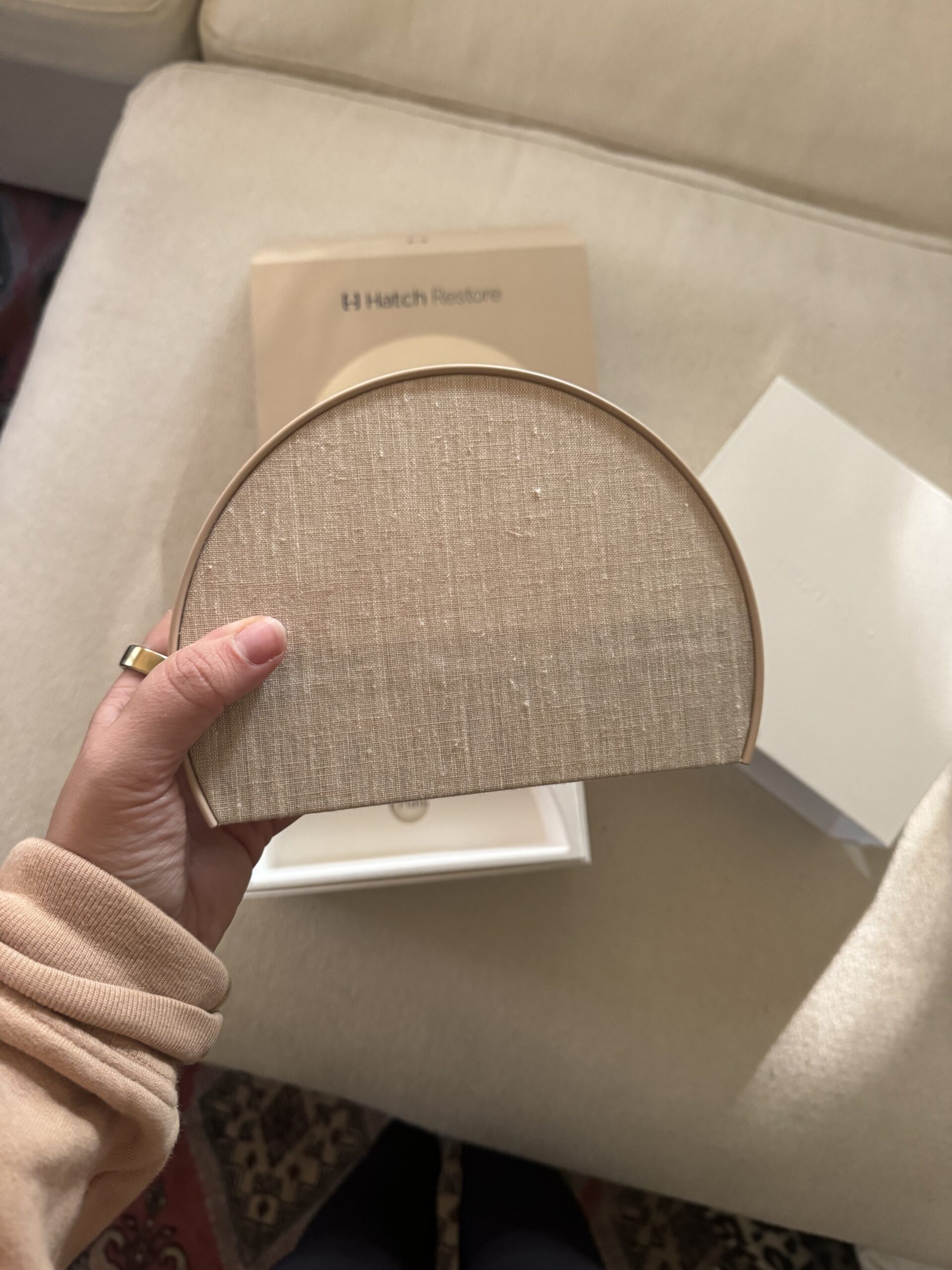 A person holds a semi-circular beige fabric-covered item next to white packaging and a cardboard box labeled "Hatch Restore" on a light-colored sofa.