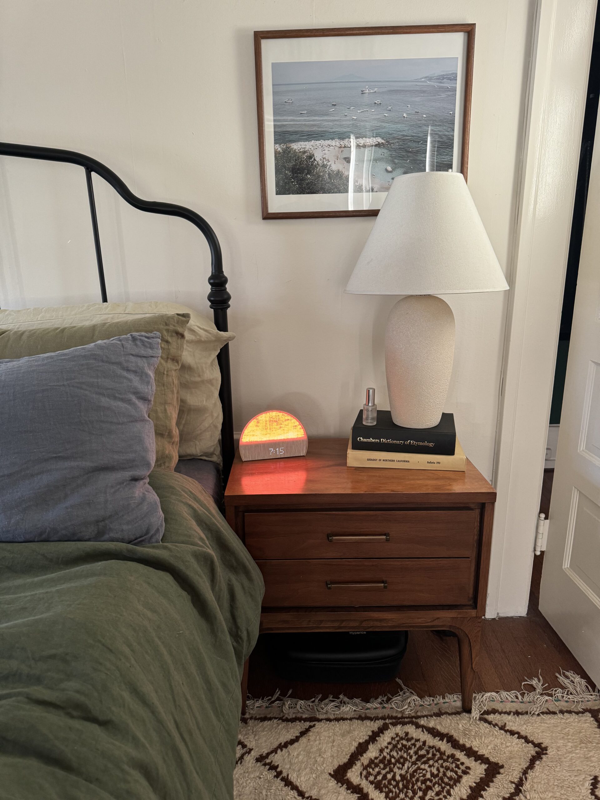 A bedside table with a lamp, clock, and book. The bed has green bedding and several pillows. A framed picture hangs on the wall above.