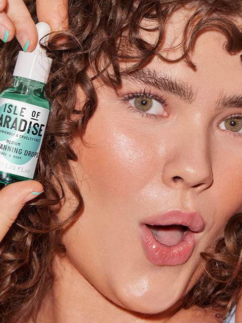 A person with curly hair holds a bottle of Isle of Paradise self-tanning drops close to their face, looking surprised.