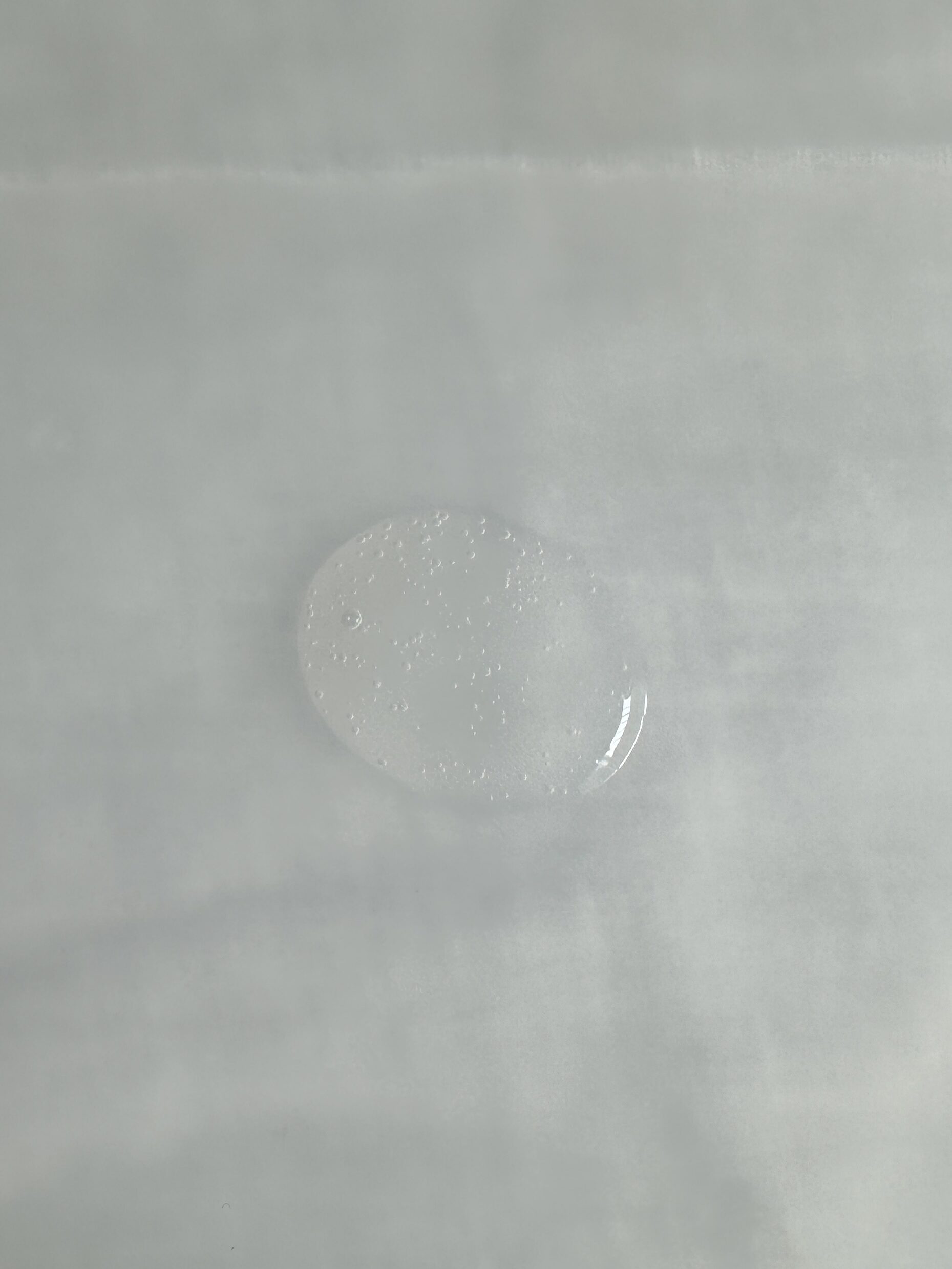 A clear, oval droplet with some small air bubbles suspended within, on a smooth, pale surface.