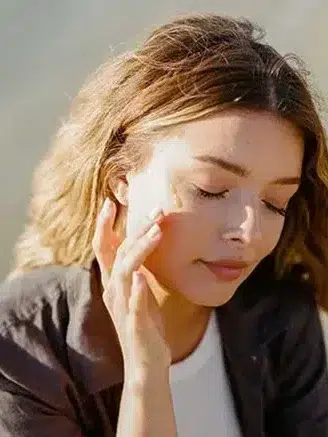 A person with closed eyes applies cream to their cheek using their fingers. They have light brown, wavy hair and are wearing a black jacket over a white top.