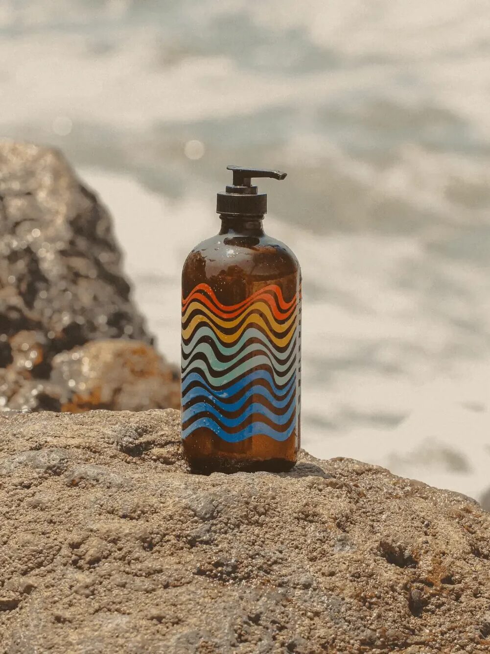 A brown bottle with a pump dispenser and wave-patterned design stands on a rock near the ocean.