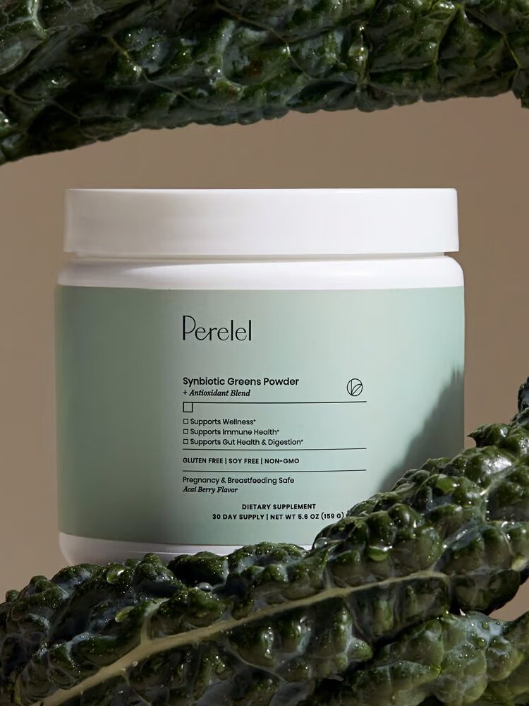 A jar of Perelel Symbiota Greens Powder stands between leafy greens. The jar's label highlights its benefits for wellness, immune health, and digestion. The product is non-GMO and gluten-free.
