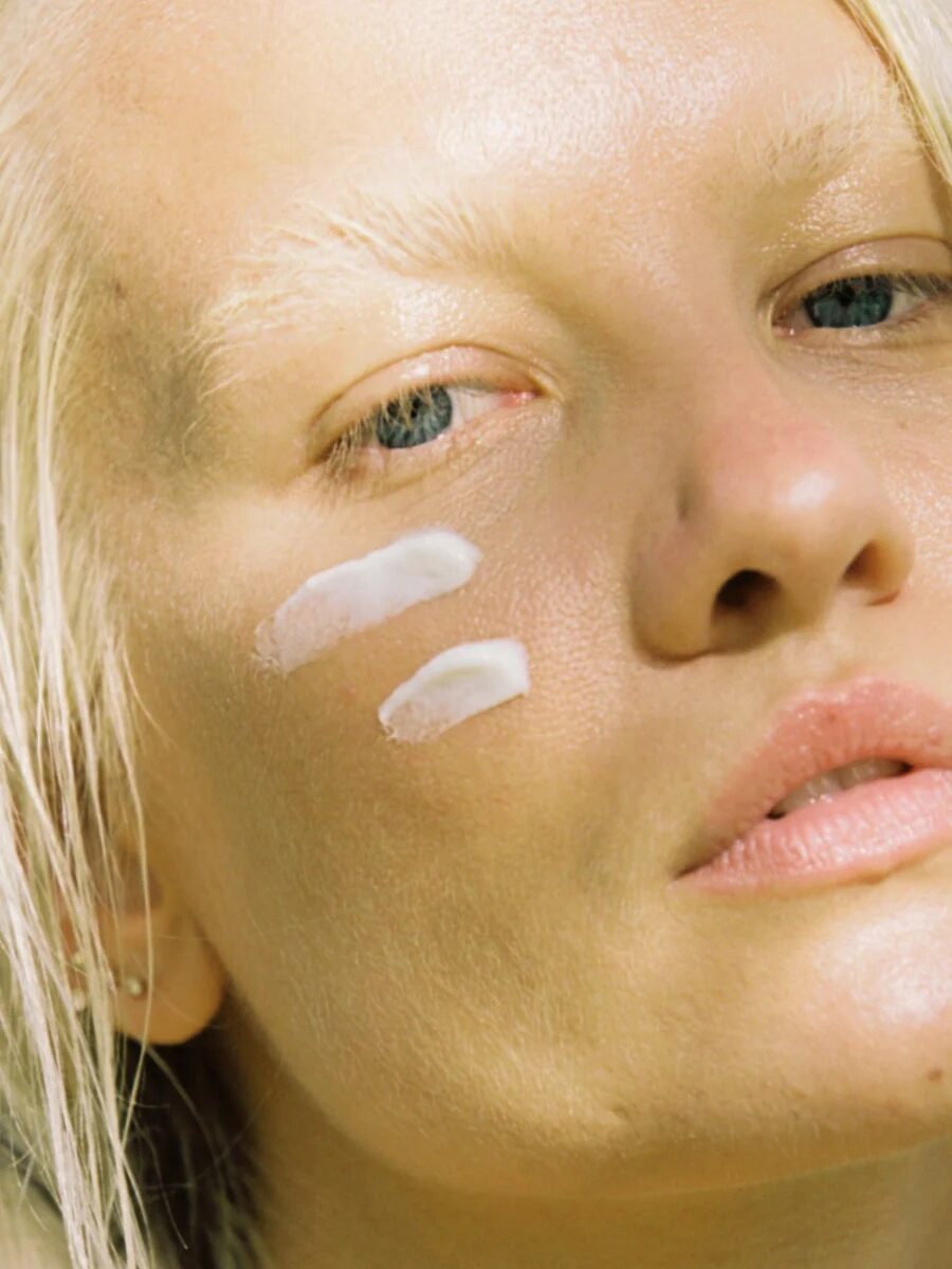 Close-up of a person's face with two streaks of white cream applied to their cheek. The person has light blonde hair and appears to be in natural lighting.