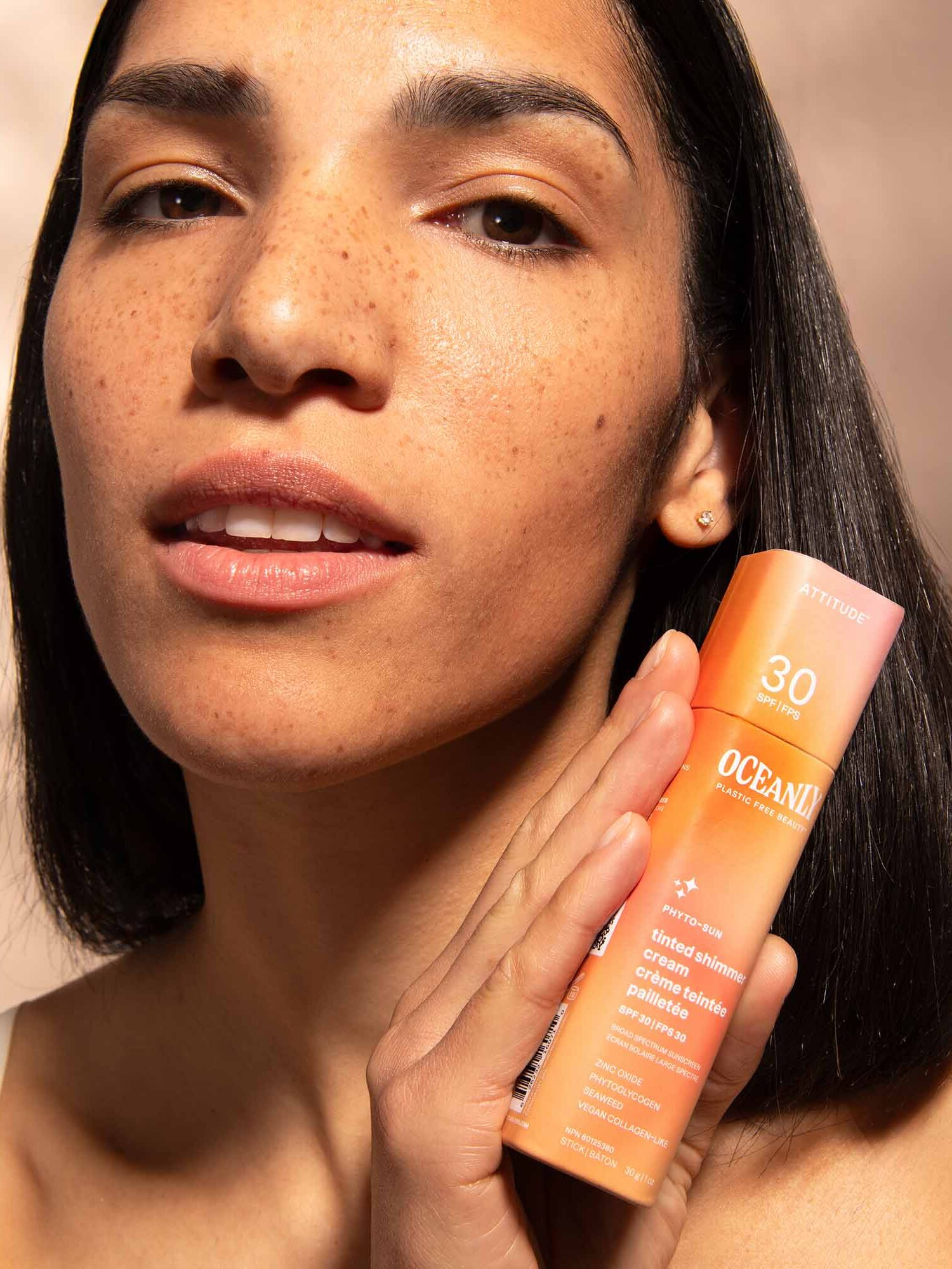 A person with long dark hair holds up a bottle of sunscreen labeled "Oceani" with SPF 30, while looking towards the camera.