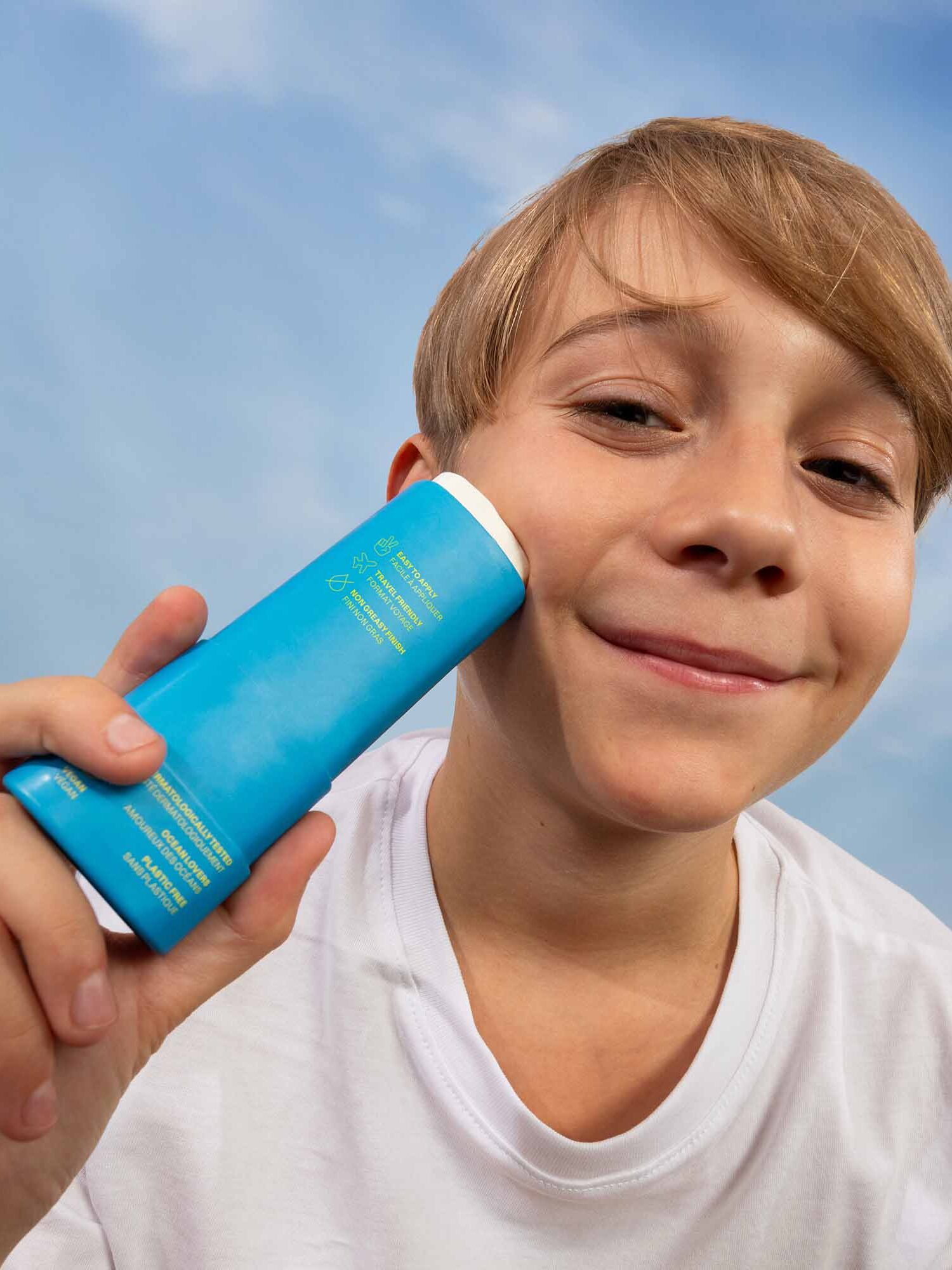 A boy wearing a white shirt holds up a blue deodorant stick, smiling outdoors against a blue sky with white clouds.