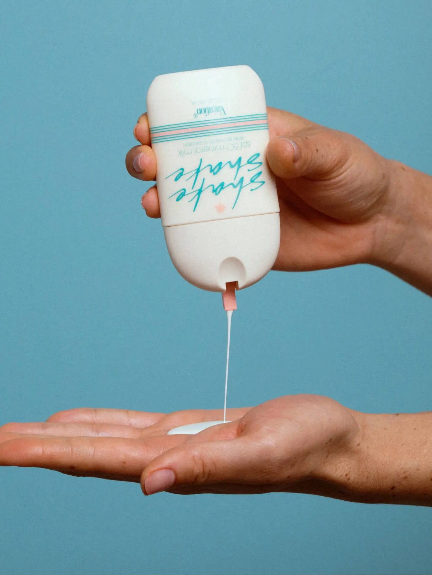 A hand is squeezing a white bottle with a blue label, dispensing a small amount of white lotion onto the palm of another hand against a blue background.