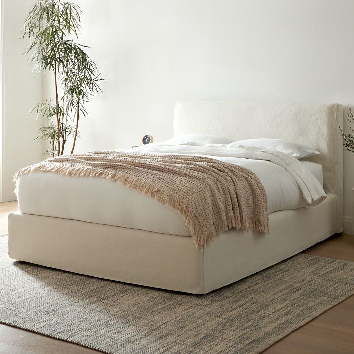 A neatly made bed with white bedding and a beige knit blanket on top stands on a light-colored area rug in a minimally decorated room with a tall plant in the corner.