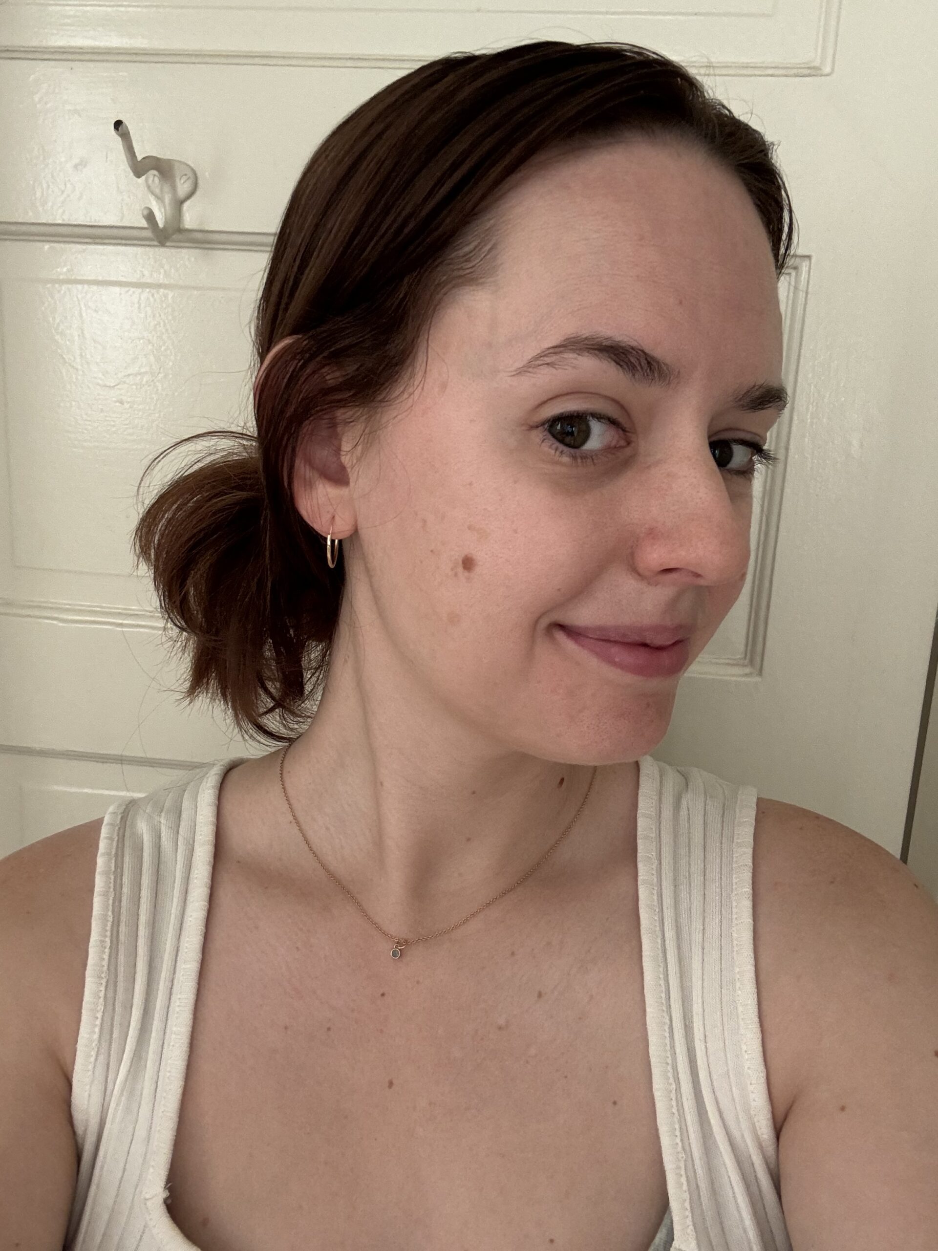 A person with brown hair tied back, wearing a white sleeveless top and a gold necklace, smiles slightly in a close-up selfie.