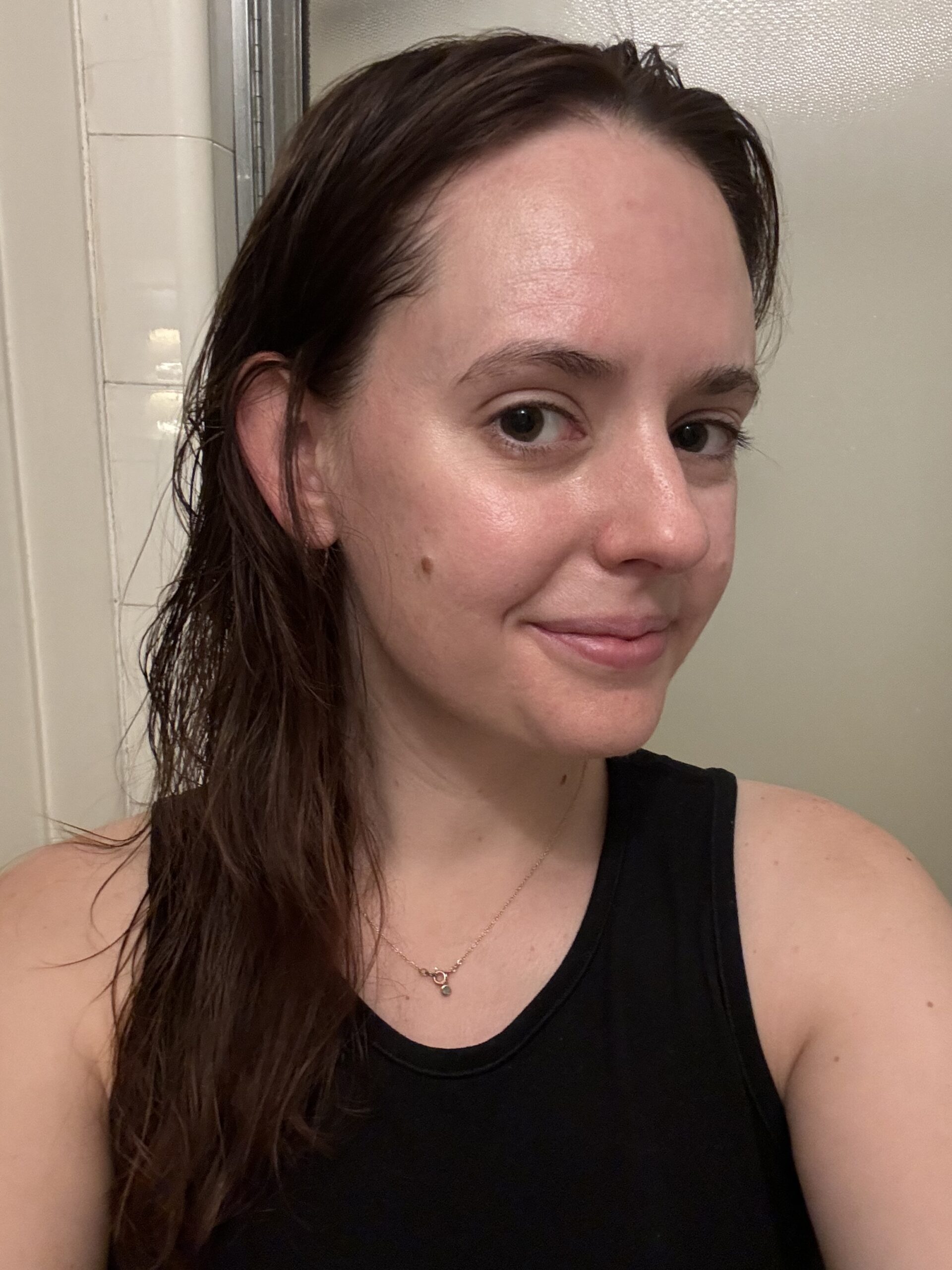 A person with wet hair, wearing a black sleeveless top, smiles at the camera. They are standing indoors in front of a shower or bathroom wall.