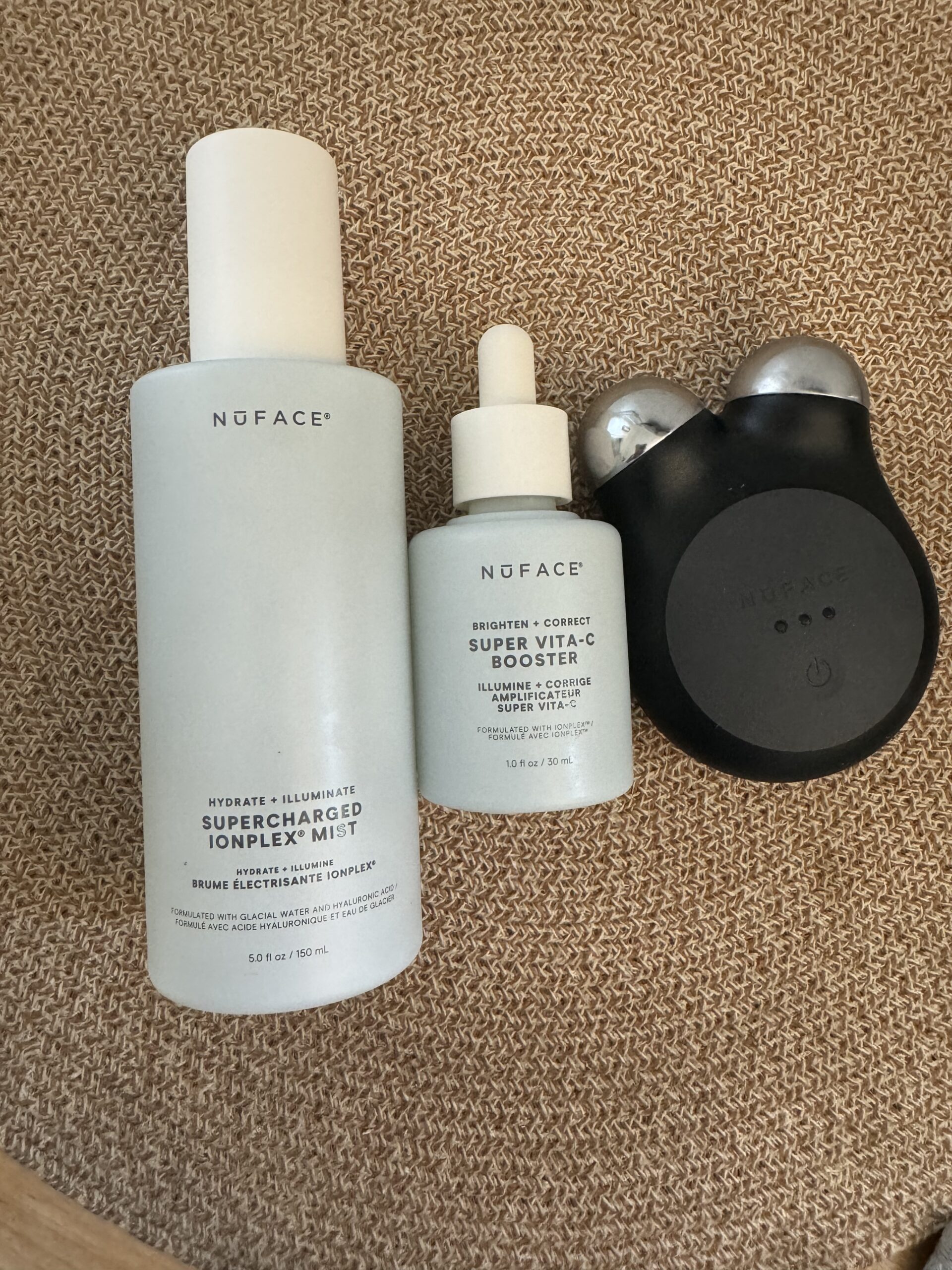 Skincare products on display: a bottle of NOFACE Supercharged IonPlex+ Mist, a bottle of NOFACE Super Vita-C Booster serum, and a black electronic facial device. They are placed on a textured surface.