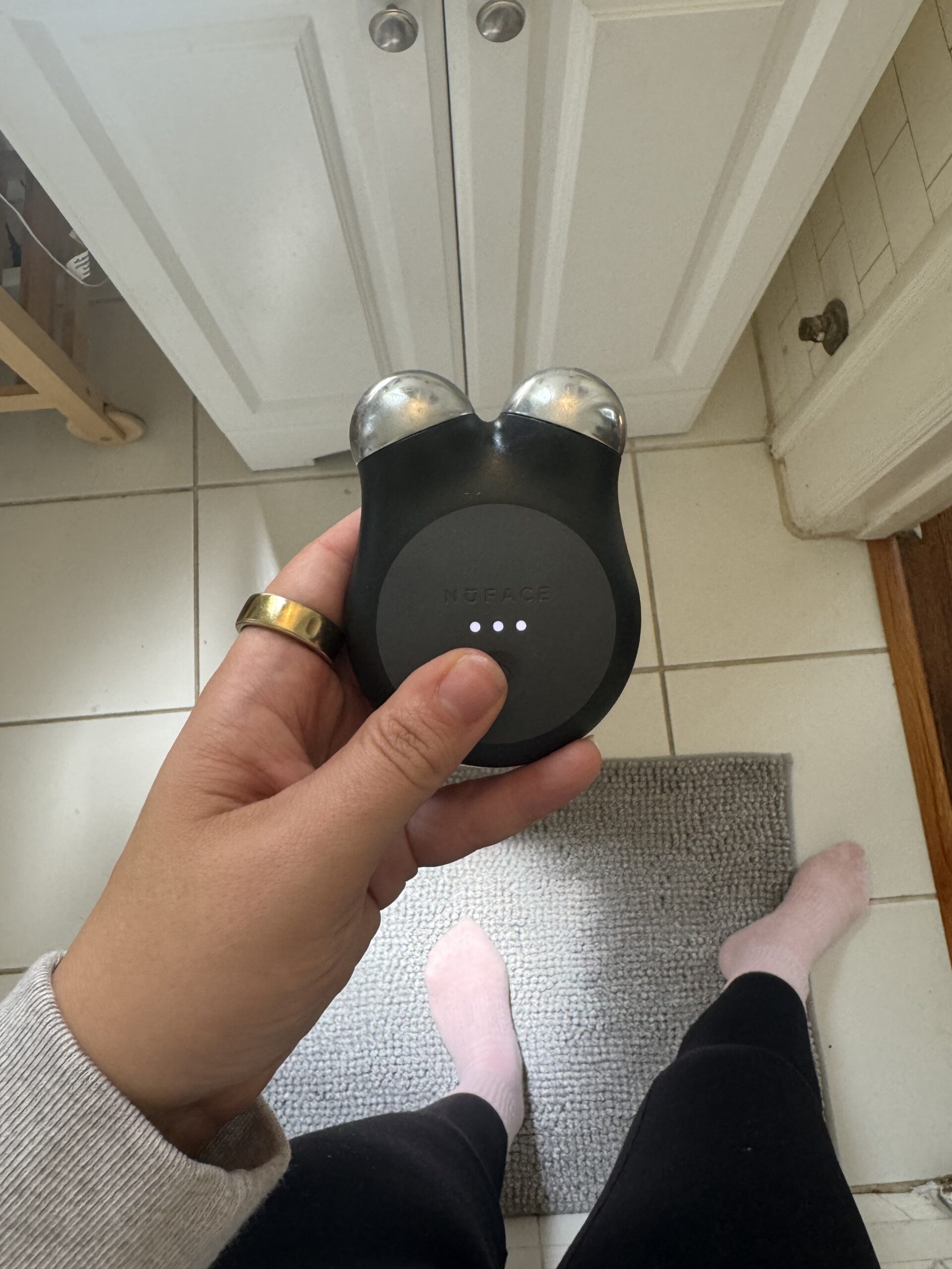 A person holds a black microcurrent facial device above a gray rug in a white-tiled room.