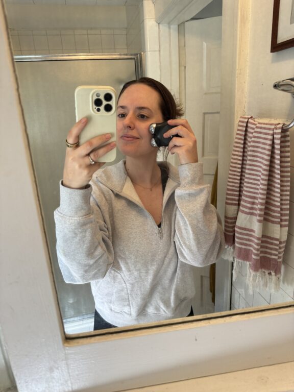 A person stands in a bathroom, holding a phone with a white case to take a selfie while using a shaving device on their neck with their other hand. They wear a light grey sweatshirt.