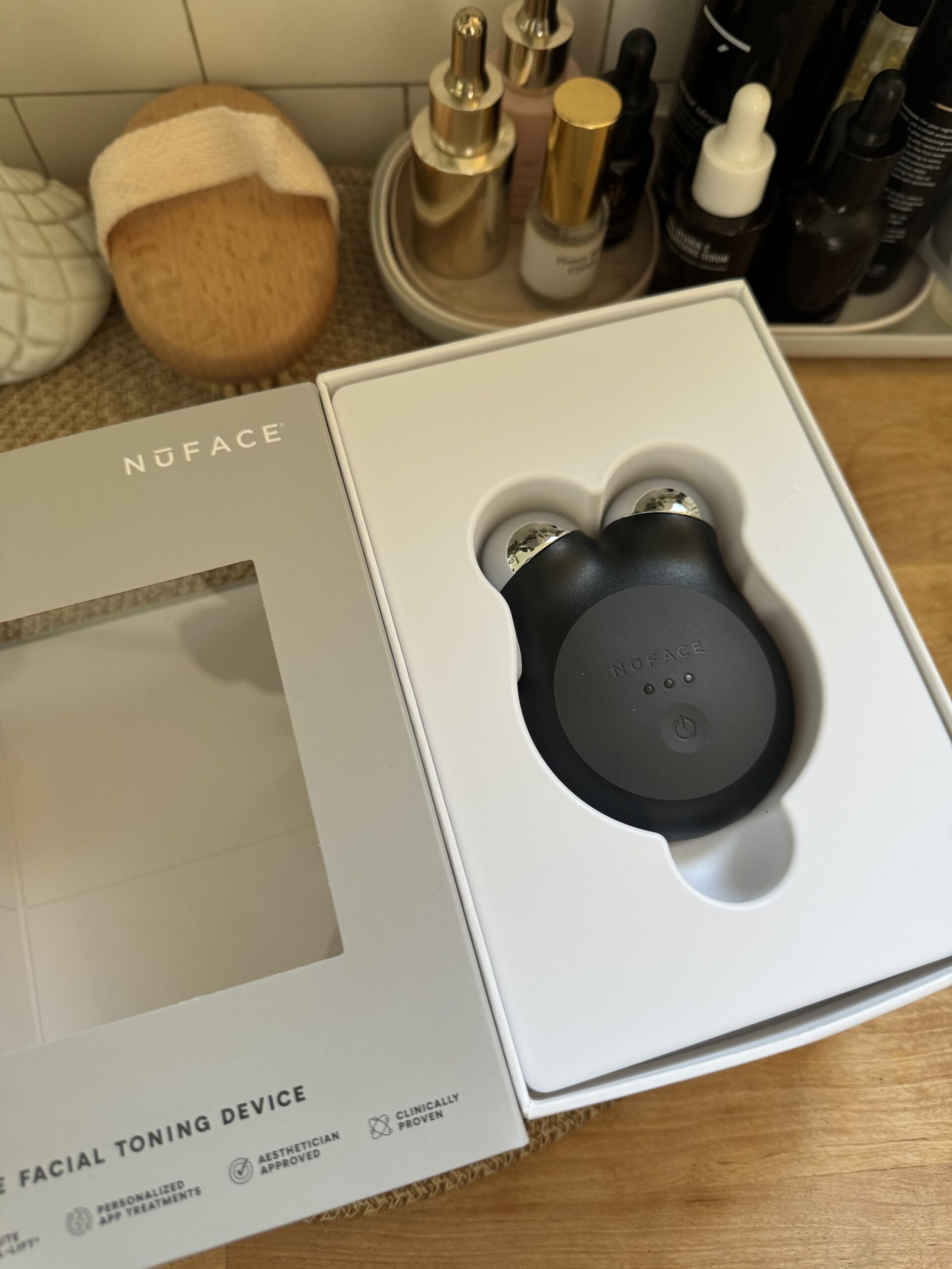 NuFACE facial toning device in its original packaging, placed on a wooden surface with various skincare products in the background.