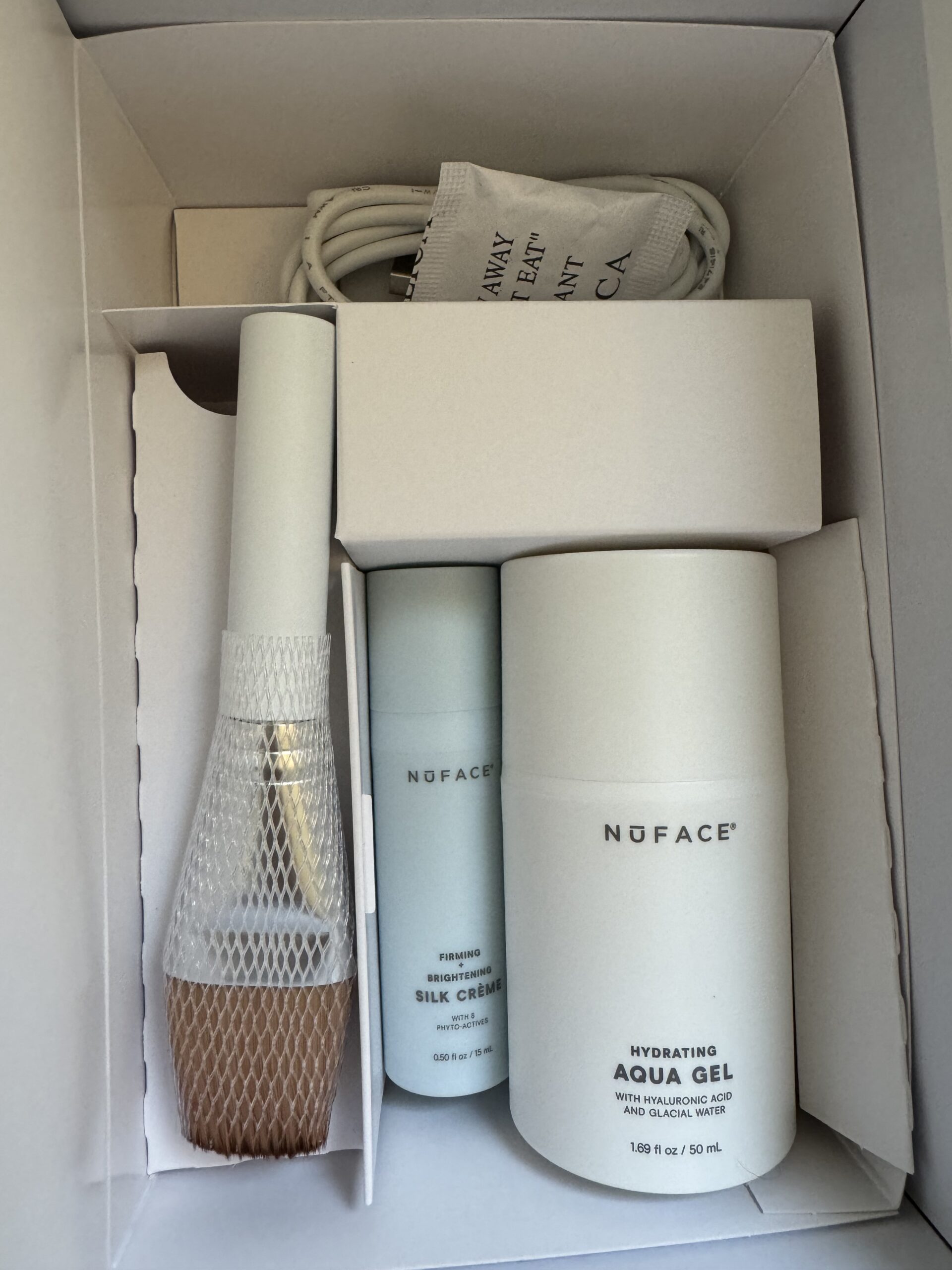 A box containing a brush with netting, a small bottle labeled "Silk Crème," a larger bottle labeled "Aqua Gel," and a coiled white power cable.