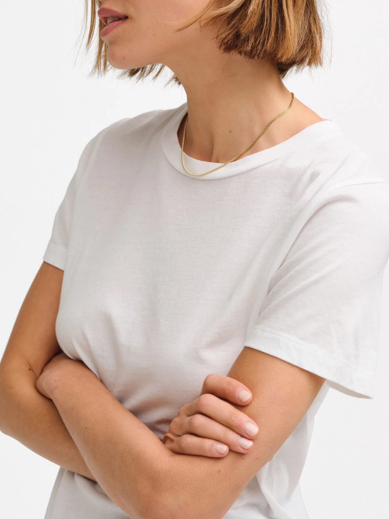 A person with short brown hair wearing a plain white t-shirt and a gold necklace, standing with arms crossed against a white background.