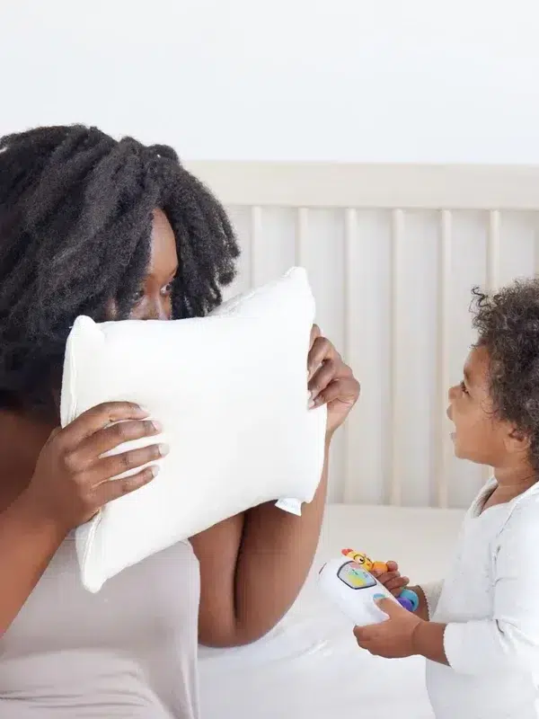 An adult holds a white pillow up to their face while a young child, holding a colorful toy, looks up at them. They are indoors, likely in a bedroom with a white background.