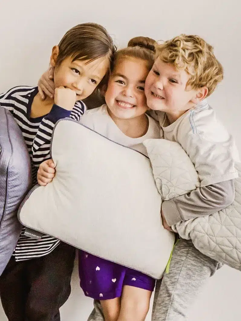 Three children standing together, smiling and holding pillows. The child on the left is wearing a striped shirt, the middle child is in a white shirt and purple shorts, and the child on the right is in a grey shirt.