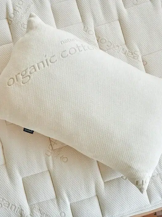 A beige pillow made of organic cotton rests on a quilted mattress with a similar organic cotton texture. Both items have the text "organic cotton" embroidered on them.