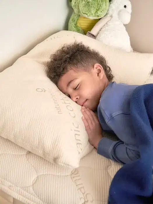 A child with curly hair sleeps peacefully on a beige bed, hugging a pillow and covered with a blue blanket. Stuffed toys are placed in the background.