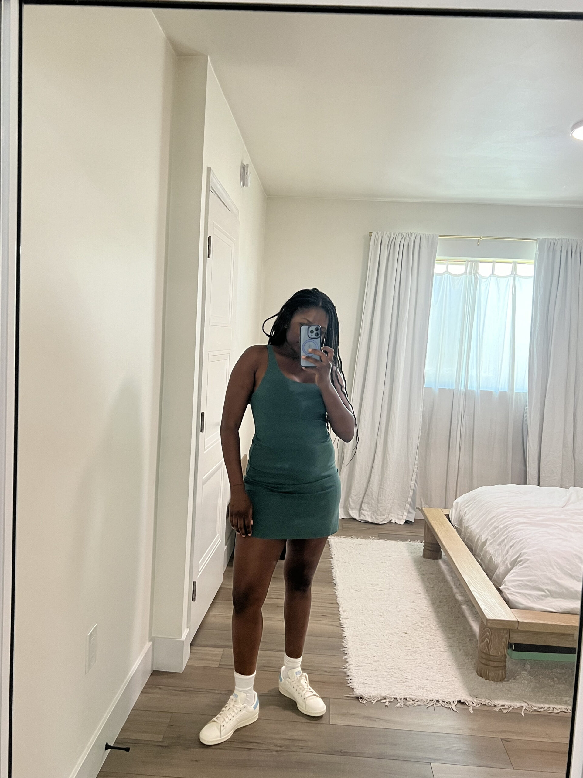 A person in a green dress takes a mirror selfie in a minimalist bedroom. The person is wearing white sneakers. The room has a wooden bed, white curtains, and a white rug.
