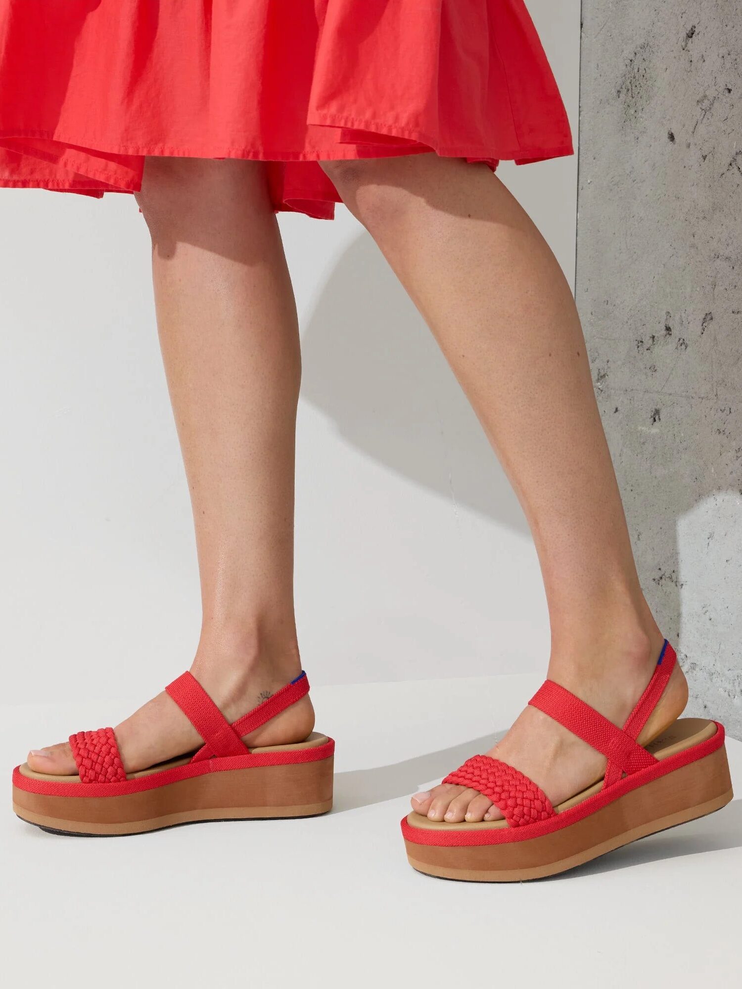 A person wearing red sandals and a red skirt stands on a concrete surface. The sandals have a thick wooden sole and woven straps.