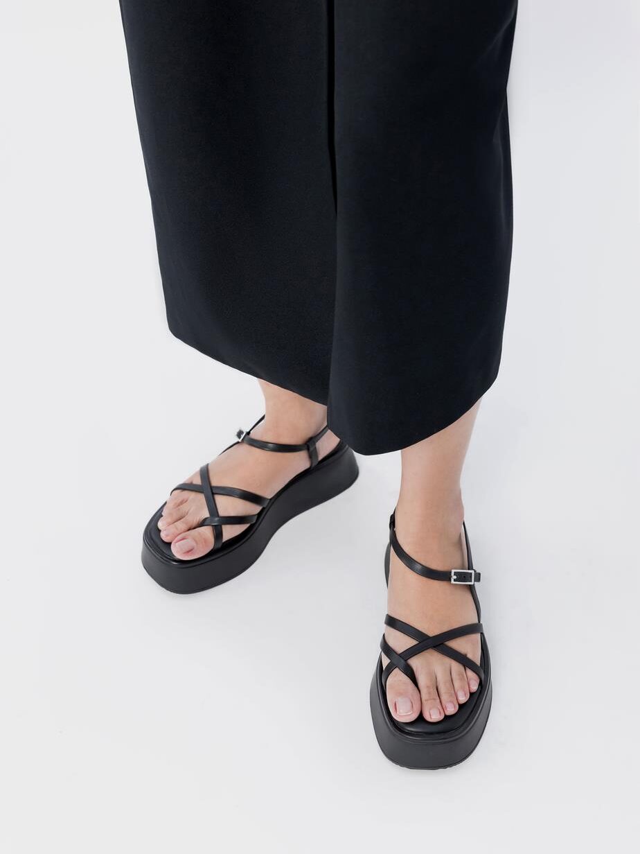 Person wearing black sandals with crisscross straps and black pants standing on a white background.