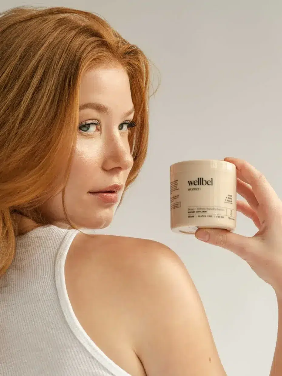 Person with long red hair in a white sleeveless top holding a beige container of Wellbel dietary supplement, facing the camera over their shoulder against a plain background.