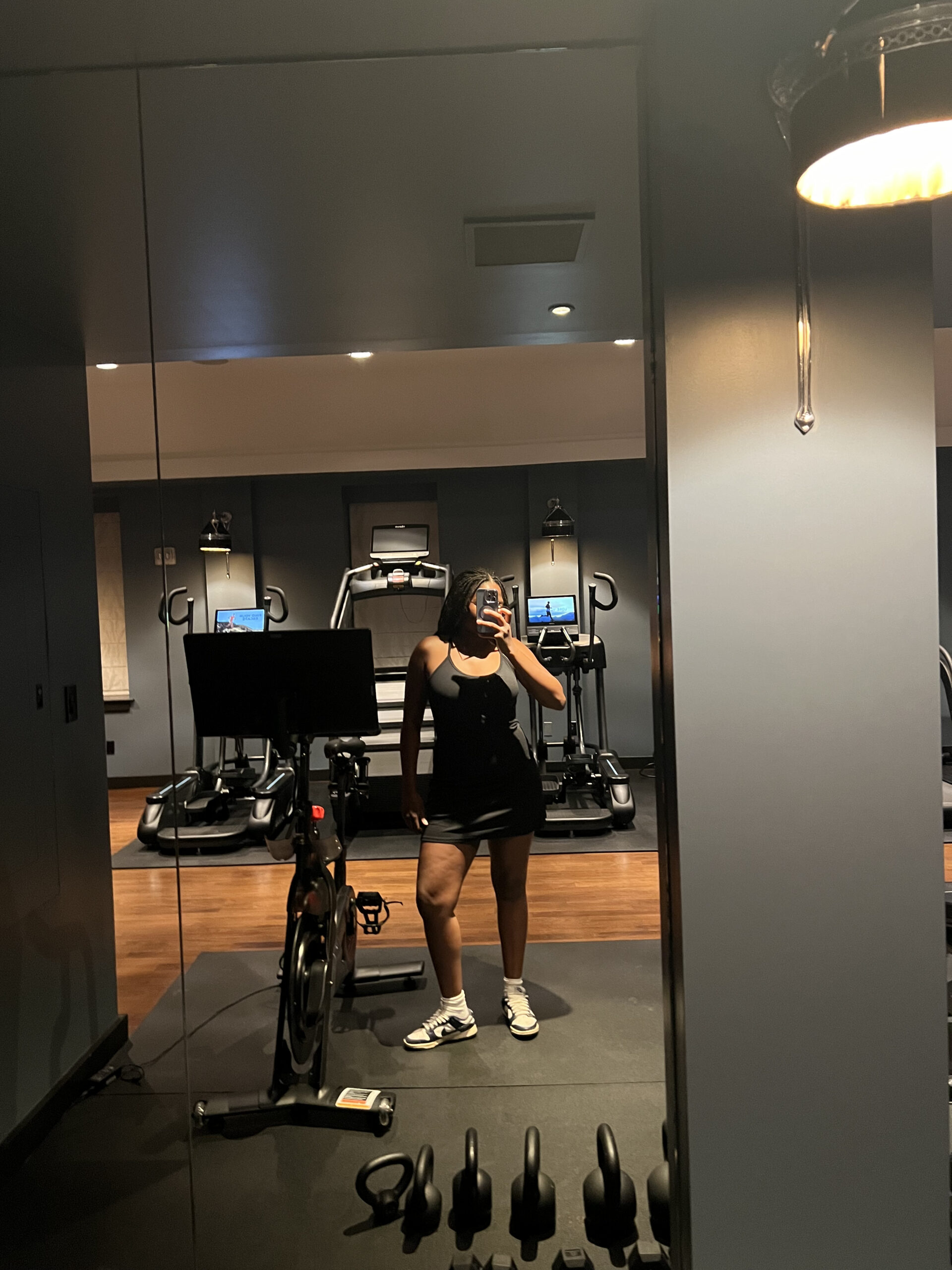 A person wearing a black workout outfit takes a mirror selfie in a gym, surrounded by exercise equipment, including stationary bikes and treadmills.