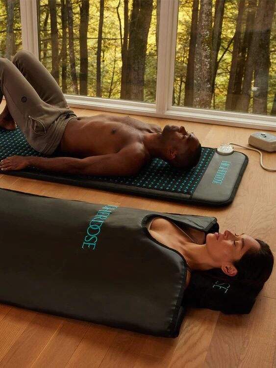 Two individuals relaxing on FAR IR mats in a room with large windows overlooking a forest. One person is lying on the mat, while the other is partially covered in a heated FAR INFRARED sleeping bag.