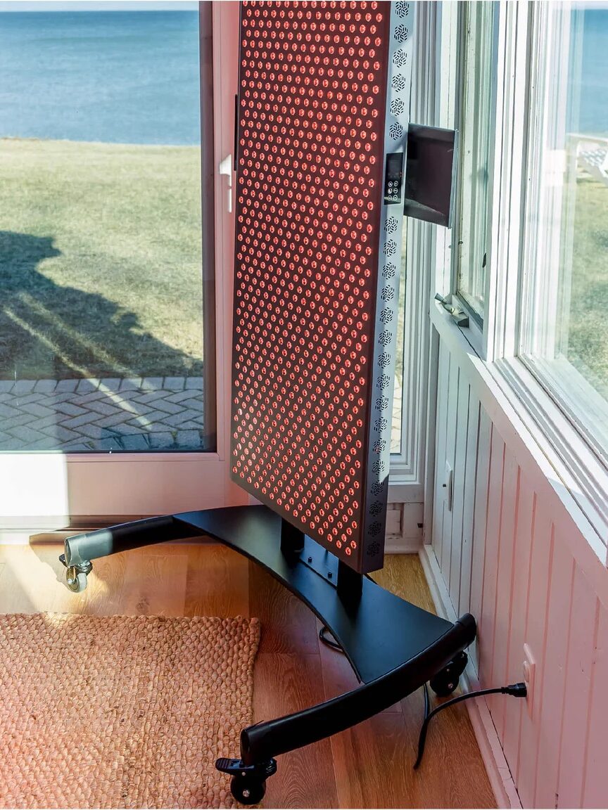 A red light therapy panel on a wheeled stand is positioned by windows overlooking a grassy area and a body of water.