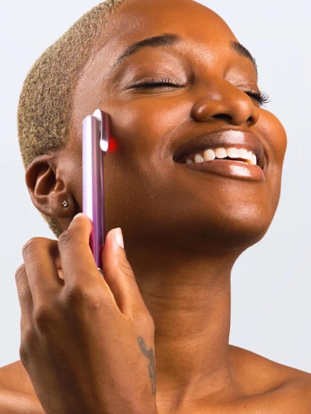 A person with short hair is smiling with eyes closed while using a purple handheld device on their cheek.