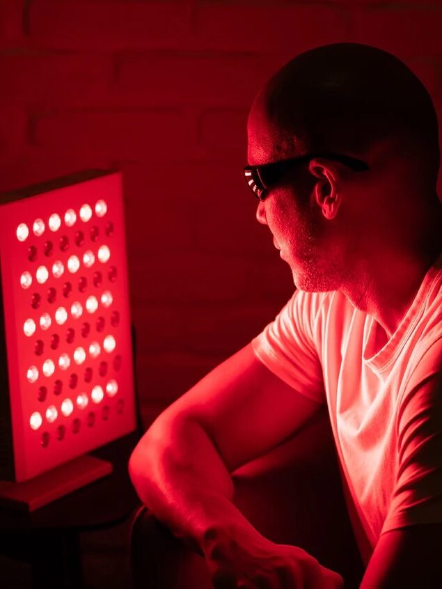 A person in a white shirt and dark glasses sits next to a panel emitting red light.