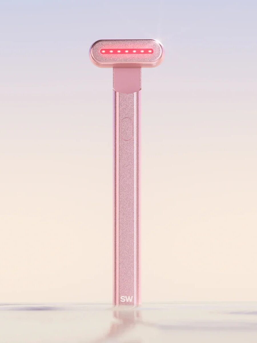 A pink skincare device with red LED lights and a long handle stands upright against a gradient background.